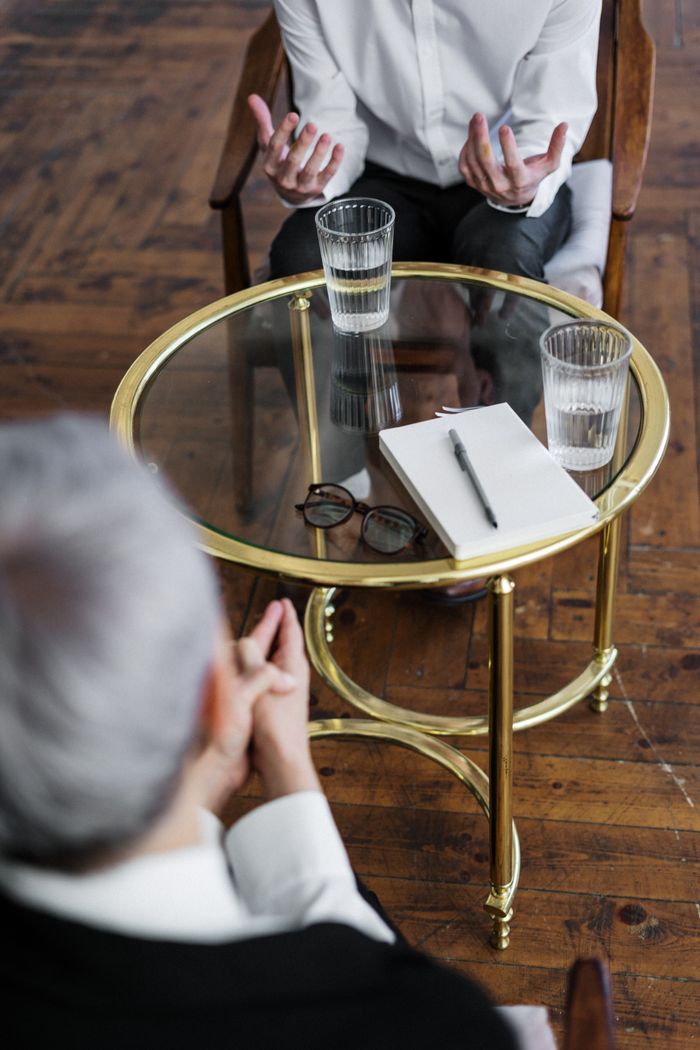services-02 Two adults discussing mental health in a counseling session across a glass table indoors.